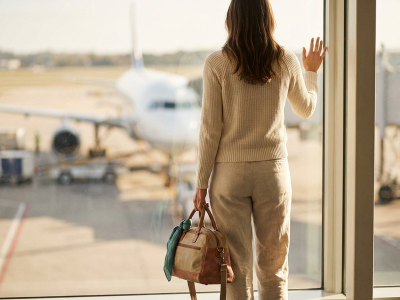 Woman with a small carry-on standing at an airport window, watching a plane and steadying herself before boarding