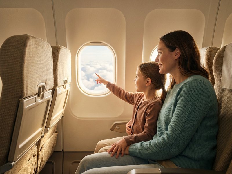 A mother and her young daughter in an airplane window seat, the girl pointing at clouds outside the window